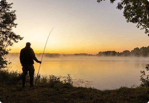 Angler steht bei Sonnenaufgang mit seiner Rute am nebligen Seeufer und blickt ruhig über das Wasser