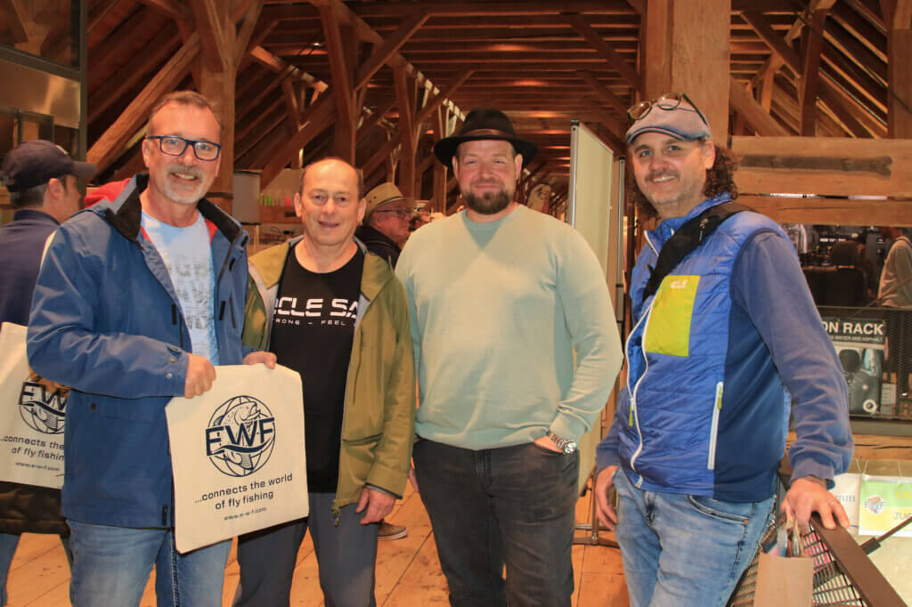 Vier Vereinsmitglieder auf der Messe Erlebniswelt Fliegenfischen in Fürstenfeldbruck, einer hält eine EWF-Stofftasche, im Hintergrund der historische Dachstuhl